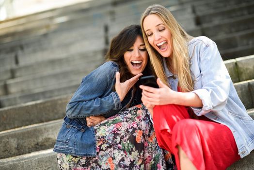 Two young women looking at some funny thing on their smart phone outdoors, sitting on urban steps. Friends girls laughing.