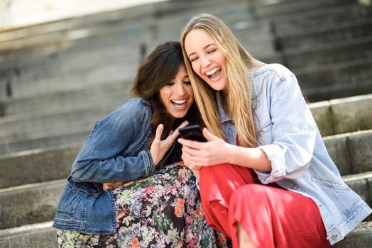 Two girls looking at some funny video on their smart phone outdoors, sitting on urban steps. Female friends girls laughing.