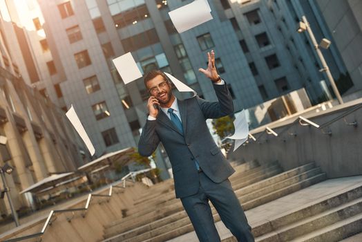 Success Portrait of happy bearded businessman in full suit talking by phone with client and tossing documents into the air while standing outdoors. Business concept
