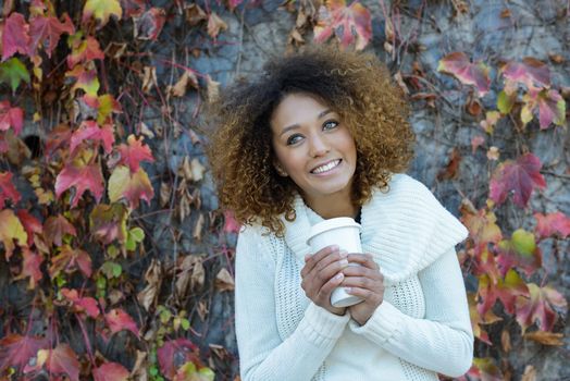 Beautiful young African American woman with afro hairstyle and green eyes wearing white winter dress. Girl drinking coffee in park sitting on grass wearing casual clothes smiling.