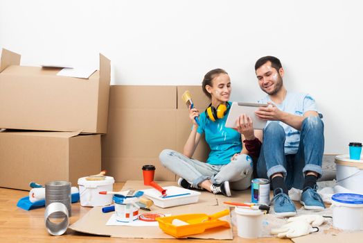 Happy couple sitting on floor and looking at tablet computer screen. Home remodeling and house interior redesign. Painting tools and materials on floor. Young family renovating their home after moving