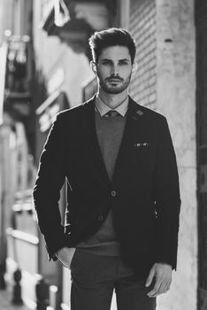 Attractive man in the street wearing british elegant suit. Young bearded businessman with modern hairstyle in urban background. Black and white photograph.