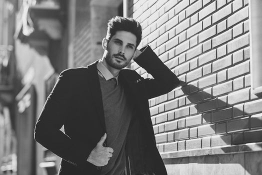 Attractive man in the street wearing british elegant suit. Young bearded businessman with modern hairstyle in urban background. Black and white photograph.
