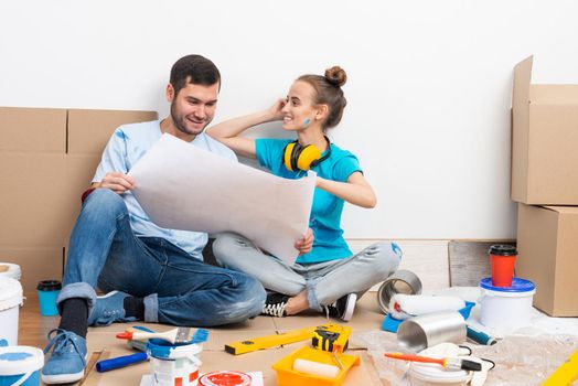 Young man and woman together planning their home renovation. Cardboard boxes, painting tools and materials on floor. House remodeling and interior renovation. People looking at blueprint at home.