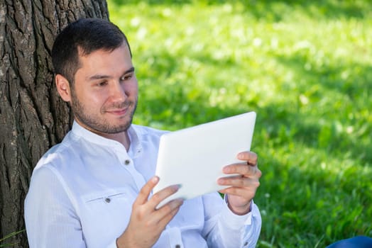 Man using tablet computer under tree in park on sunny day. Handsome guy looking at tablet screen. Happy smiling man sitting on green grass leaning against tree. Digital technology and communication