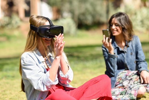 Woman looking in VR glasses and gesturing with his hands outdoors in urban park with her female friend taking a video with smart phone.
