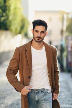 Young man wearing winter clothes in the street. Young bearded guy with modern hairstyle with coat, blue jeans and white t-shirt.