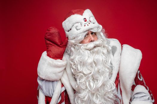 Stock photo of white-bearded Father Frost listening attentively to something with hand by ear. Looking at camera with doubt face.