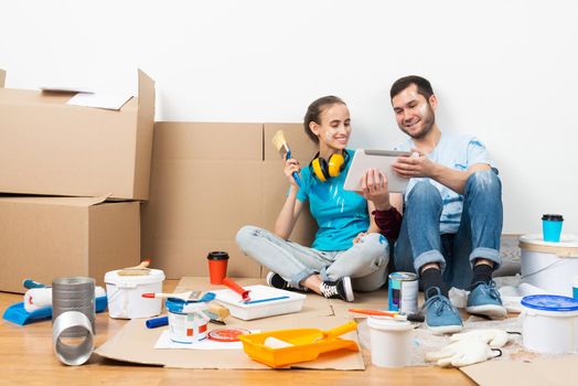 Happy couple watching something funny on tablet computer while sitting on floor. Painting tools and materials for home remodeling and interior redesign. Young family renovating their home after moving