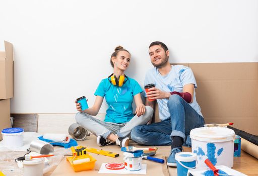 Happy smiling couple relaxing on floor with cup of coffee among construction tools and materials. House remodeling and interior renovation concept. Young man and woman together painting wall at home.