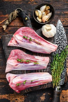 Raw lamb shanks meat on a cutting board with herbs. Dark wooden background. Top view.