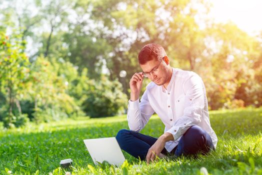 Young businessman sitting on green grass and using laptop computer. Handsome man working with computer in park at sunny summer day. Outdoors nature journey and relaxation. Freelance work concept.