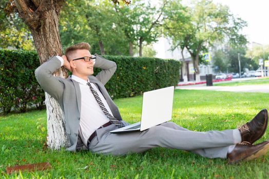 a young businessman in the park with a laptop, resting near a tree. work out of the office