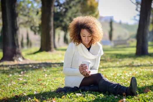 Beautiful young African American woman with afro hairstyle and green eyes wearing white winter dress. Girl drinking coffee in park sitting on grass wearing casual clothes smiling.