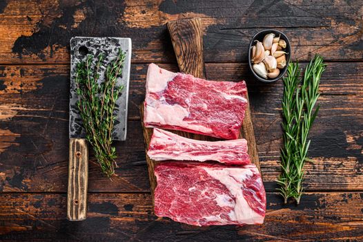 Raw beef calf short ribs meat on a butcher cutting board with cleaver. Dark wooden background. Top view.