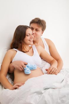 happy young pregnant woman with husband on white bed in light bedroom with baby booties