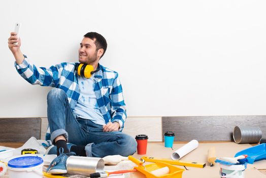 Smiling caucasian man taking selfies photo on mobile phone while sitting near wall. House remodeling and interior renovation. Young bearded guy sitting on floor among construction tools at home.