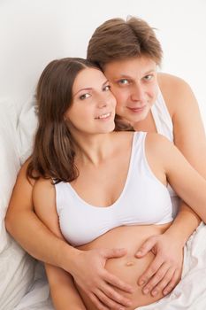 happy young pregnant woman with husband on white bed in light bedroom