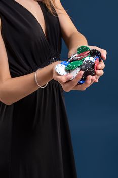 Elegant female casino player holding a handful of chips on dark blue background, hands close up. A woman in a black dress with poker chips in her hands