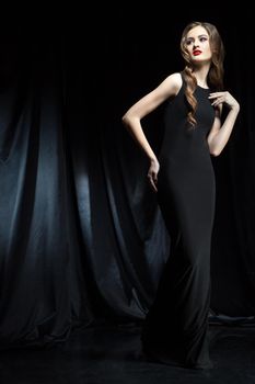 Portrait of young beautiful woman with long wavy hair in black dress posing against of black cloth