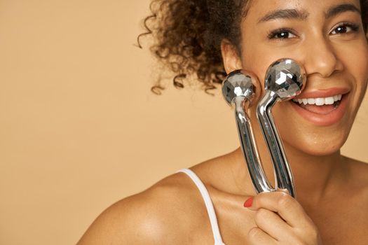 Close up portrait of adorable young woman smiling at camera while using silver metal face roller, posing isolated over beige background. Facial massage concept