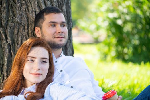 Young couple relaxing with coffee under tree in park on sunny day. Happy couple in love spend time outdoors together. Handsome man and pretty redhead girl sitting on green grass leaning against tree