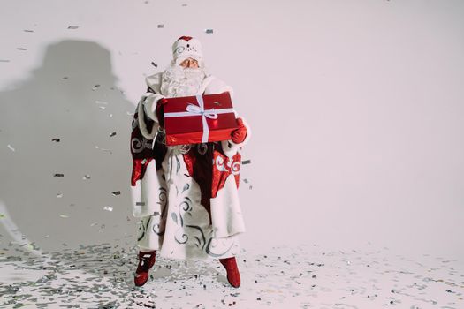 Stock photo of jolly Father Frost in festive traditional costume jumping with wrapped Christmas present in hands under falling silver confetti.