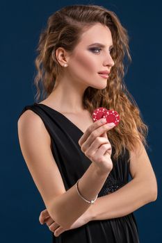 Portrait of the female gambler at the casino keeping poker chips in the hand on dark blue background. Beautiful woman in black dress holds poker chips