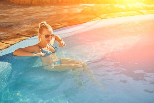 Woman in blue swimsuit and sunglasses relaxing in outdoor pool with clean transparent turquoise water. Woman sunbathing in bikini at tropical resort. Sun flare