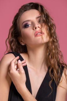 Beautiful fashionable girl with long curly hair in a black dress in the studio on a pink background. Advertising, beauty salon, cosmetics, clothing. Fashion, boutique. Pink.
