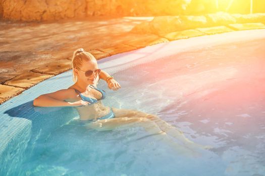 Woman in blue swimsuit and sunglasses relaxing in outdoor pool with clean transparent turquoise water. Woman sunbathing in bikini at tropical resort. Sun flare