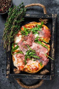 Italian Pizza with prosciutto parma ham, arugula salad and cheese in a rustic wooden tray. Black background. Top view.