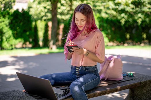 Woman writing in a notebook sitting on a wooden bench in the park. Girl working outdoors on portable computer, copy space. Technology, communication, freelance and remote working concept.