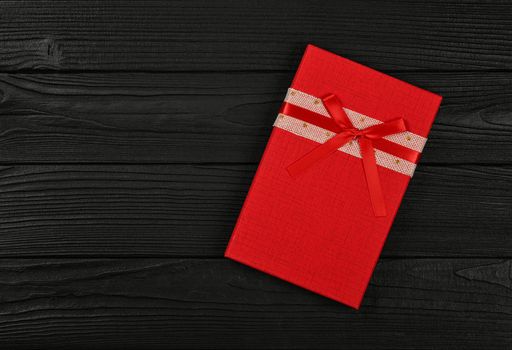 Valentine template of one closed vivid red gift box with ribbon bow over black wooden table background, close up flat lay, elevated top view, directly above