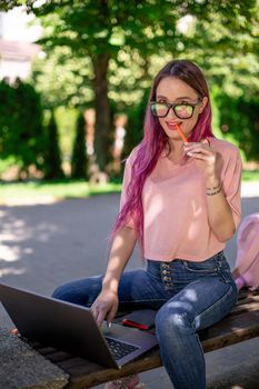 Young girl with pink hair is studying in the spring park, sitting on the wooden bench and browsing on her laptop