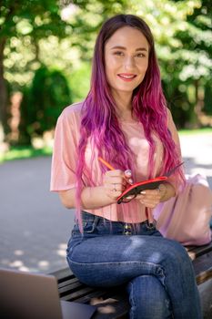 Woman writing in a notebook sitting on a wooden bench in the park. Girl working outdoors on portable computer, copy space. Technology, communication, freelance and remote working concept.