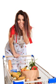 A young female pushing a shopping cart full with groceries isolated on white background. Brunette in a summer dress makes purchases. A woman laughs and closes her mouth with her palm