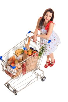 High angle view of girl smiling at camera while pushing a shopping cart full with groceries isolated on white background. Brunette in a short summer dress and red sandals