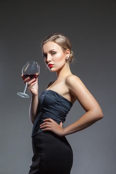 Sexual young woman posing in black dress with wine glass in hand, isolated, over the gray background.