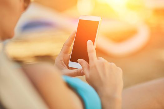 Close up view of a young attractive woman on holiday laying down on a white chaise-longue, holding and using a smartphone. Black display. People travel technology. Sun flare