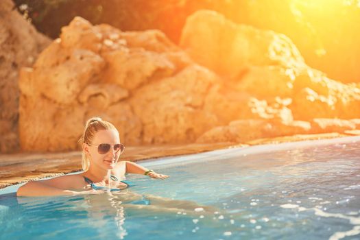 Woman in blue swimsuit and sunglasses relaxing in outdoor pool with clean transparent turquoise water. Woman sunbathing in bikini at tropical resort. Sun flare