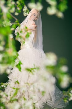 Beautiful young bride has veil over her head and face and looks at the camera. Over blurred nature background.