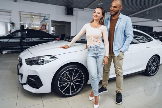 Happy couple of caucasian woman and african american man standing near their new luxury car inside car salon