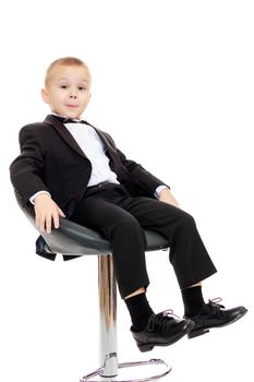 Beautiful little boy in a strict black suit , white shirt and tie.He sits in a spinning chair.Isolated on white background.