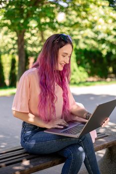 Young girl with pink hair is studying in the spring park, sitting on the wooden bench and browsing on her laptop