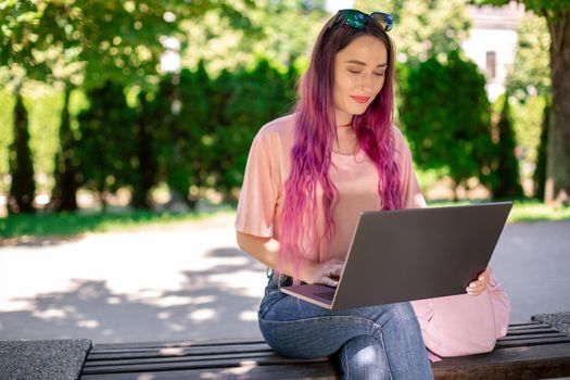 Young girl with pink hair is studying in the spring park, sitting on the wooden bench and browsing on her laptop