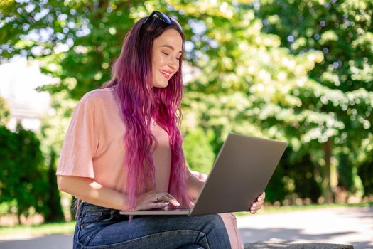 Young girl with pink hair is studying in the spring park, sitting on the wooden bench and browsing on her laptop