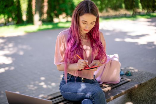 Woman writing in a notebook sitting on a wooden bench in the park. Girl working outdoors on portable computer, copy space. Technology, communication, freelance and remote working concept.