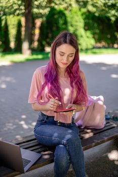 Woman writing in a notebook sitting on a wooden bench in the park. Girl working outdoors on portable computer, copy space. Technology, communication, freelance and remote working concept.