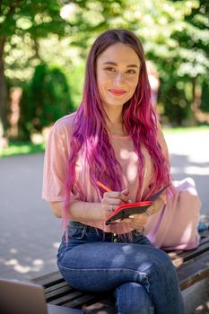 Woman writing in a notebook sitting on a wooden bench in the park. Girl working outdoors on portable computer, copy space. Technology, communication, freelance and remote working concept.